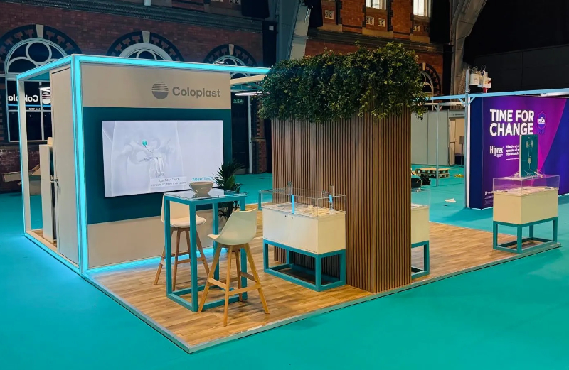Hospitality area within Coloplast’s exhibition stand, featuring wood-effect flooring, greenery, bar-height seating, AV displays, and custom-built counters.